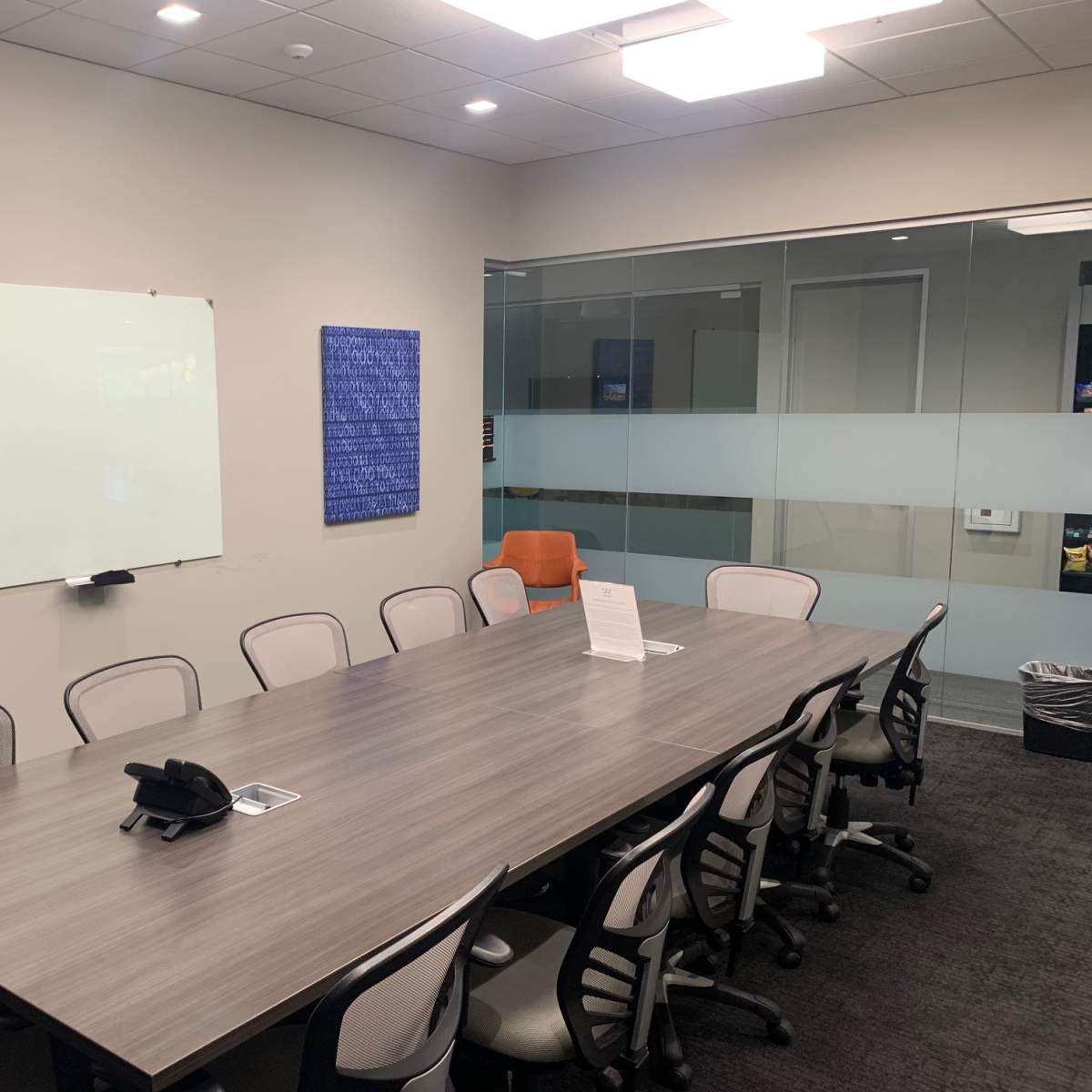 A conference room features a long table surrounded by ergonomic chairs, a whiteboard on one wall, and a glass partition revealing a workspace area.