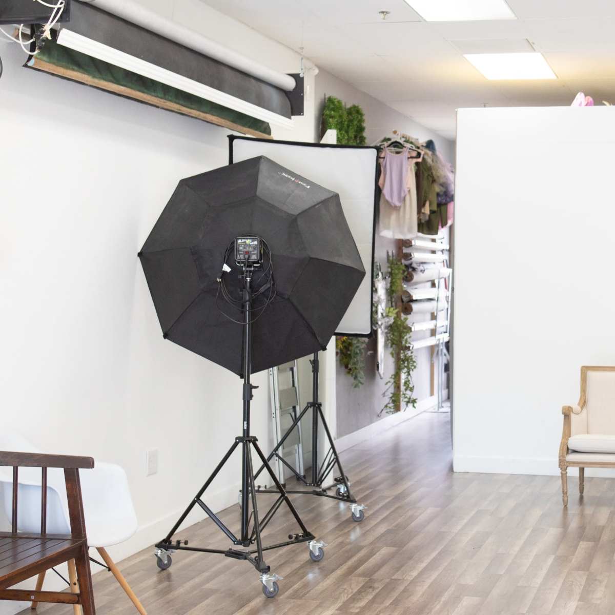 A photography studio featuring a black softbox light on a stand, a wooden bench, and a white backdrop with various props behind it.