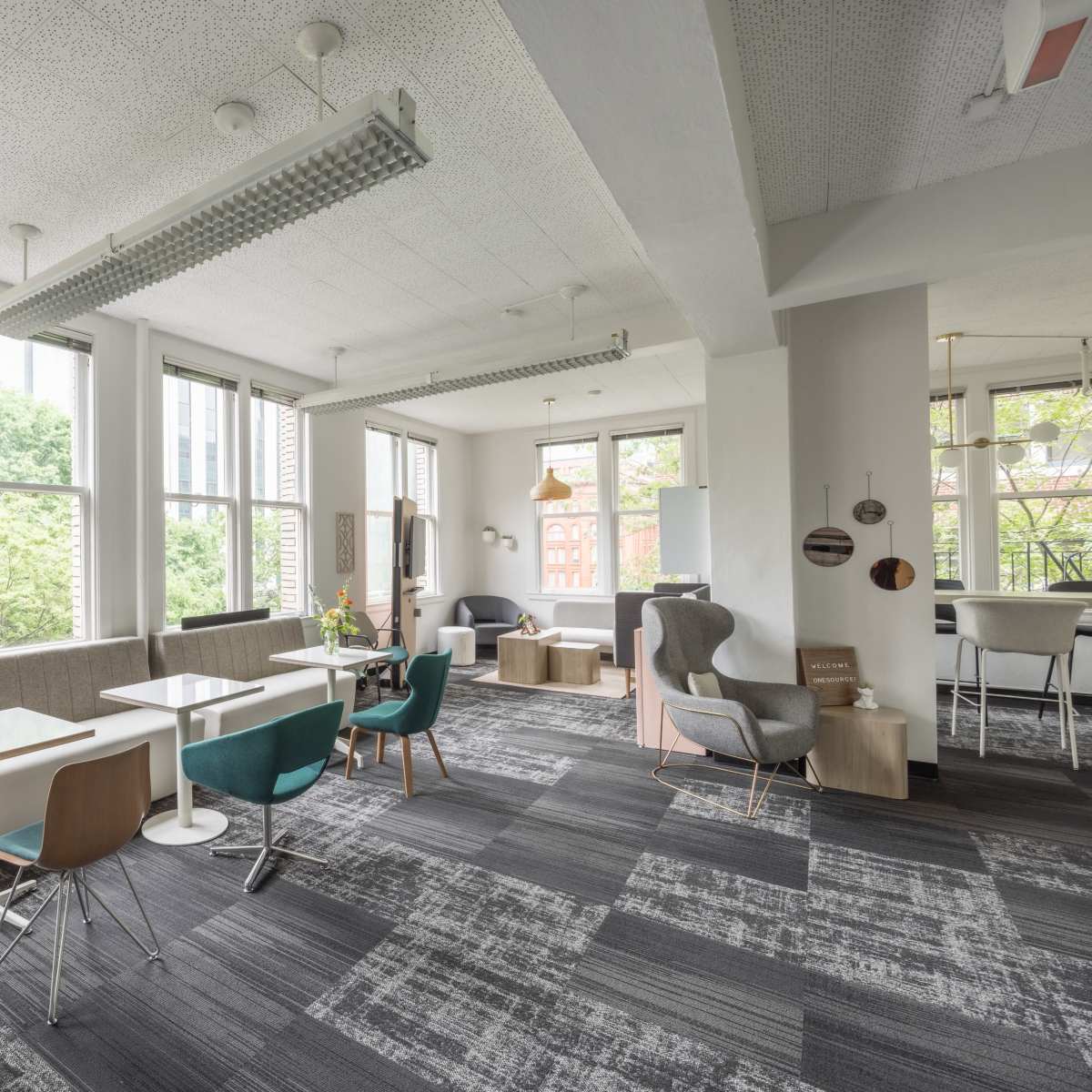 A modern workspace with several tables and chairs near large windows, featuring a mix of seating areas and greenery outside.
