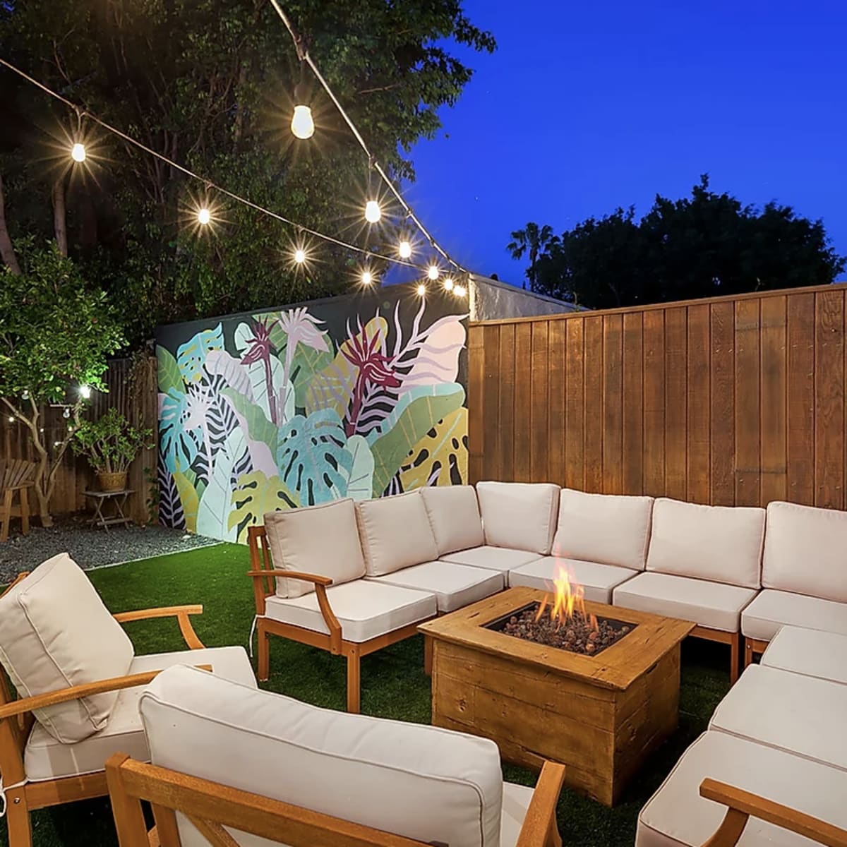 An outdoor seating area with a fire pit surrounded by a circular arrangement of cushioned chairs and a colorful mural on the wall.