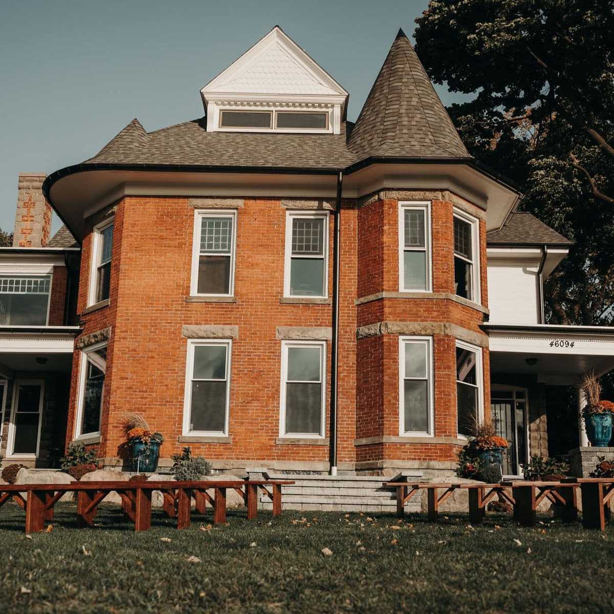 A Victorian-style brick house features a conical roof and large windows, surrounded by grass and wooden benches.