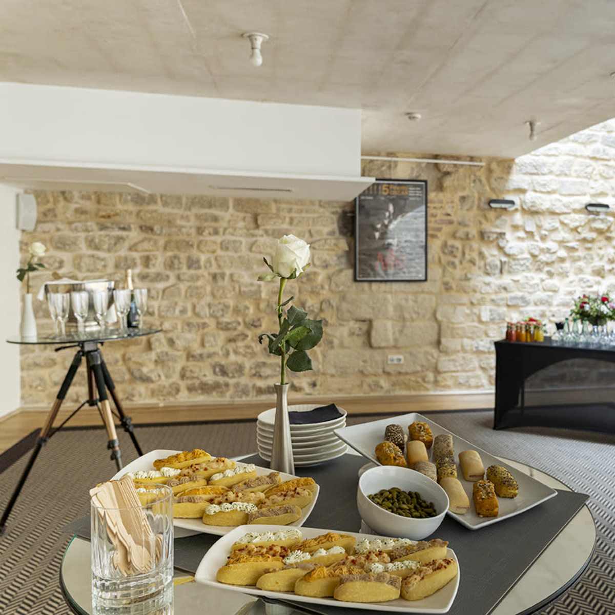A neatly arranged event space features tables with appetizers and drinks against a backdrop of stone walls and large windows.