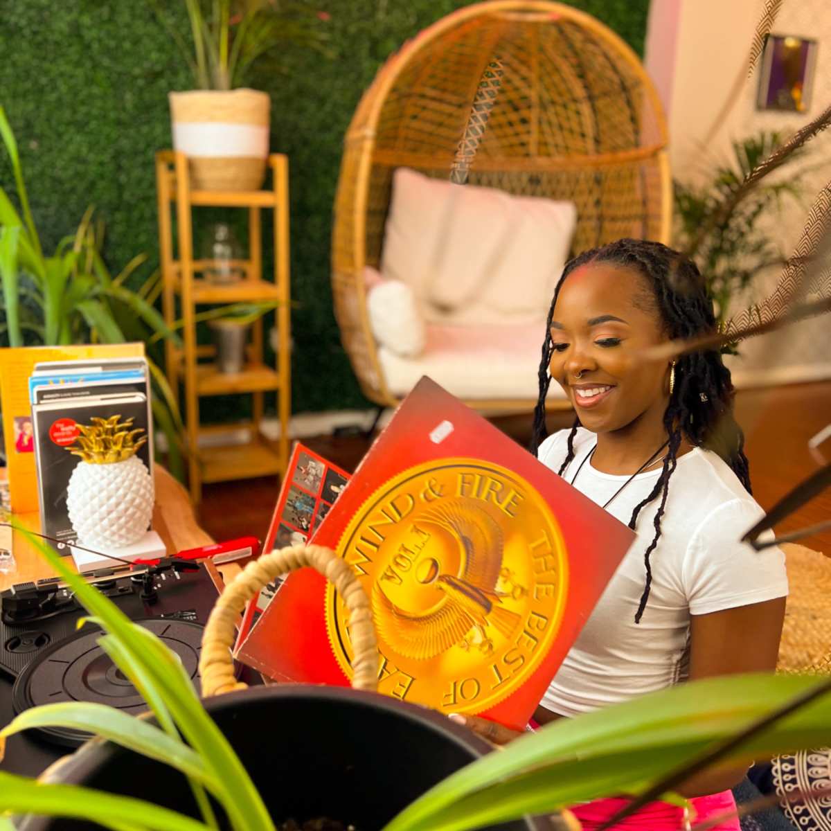 A woman sits in a decorative indoor space, looking at a vinyl record while surrounded by greenery and various decor items.