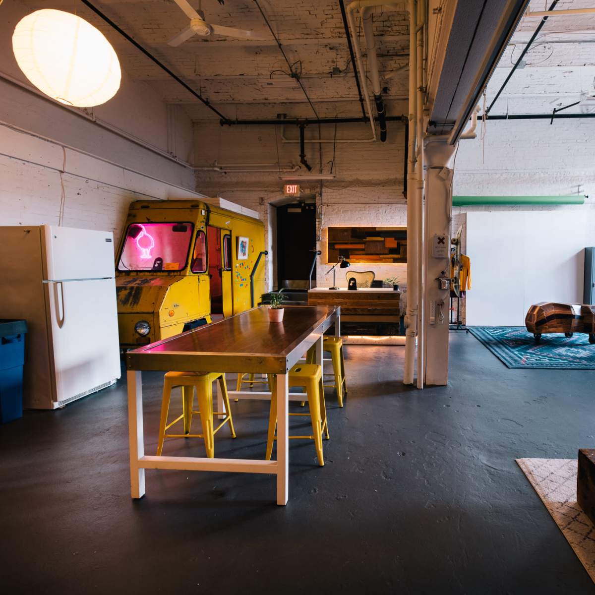 A spacious, well-lit industrial-style room features a yellow food truck, a long dining table with yellow stools, various kitchen appliances, and a relaxed seating area.