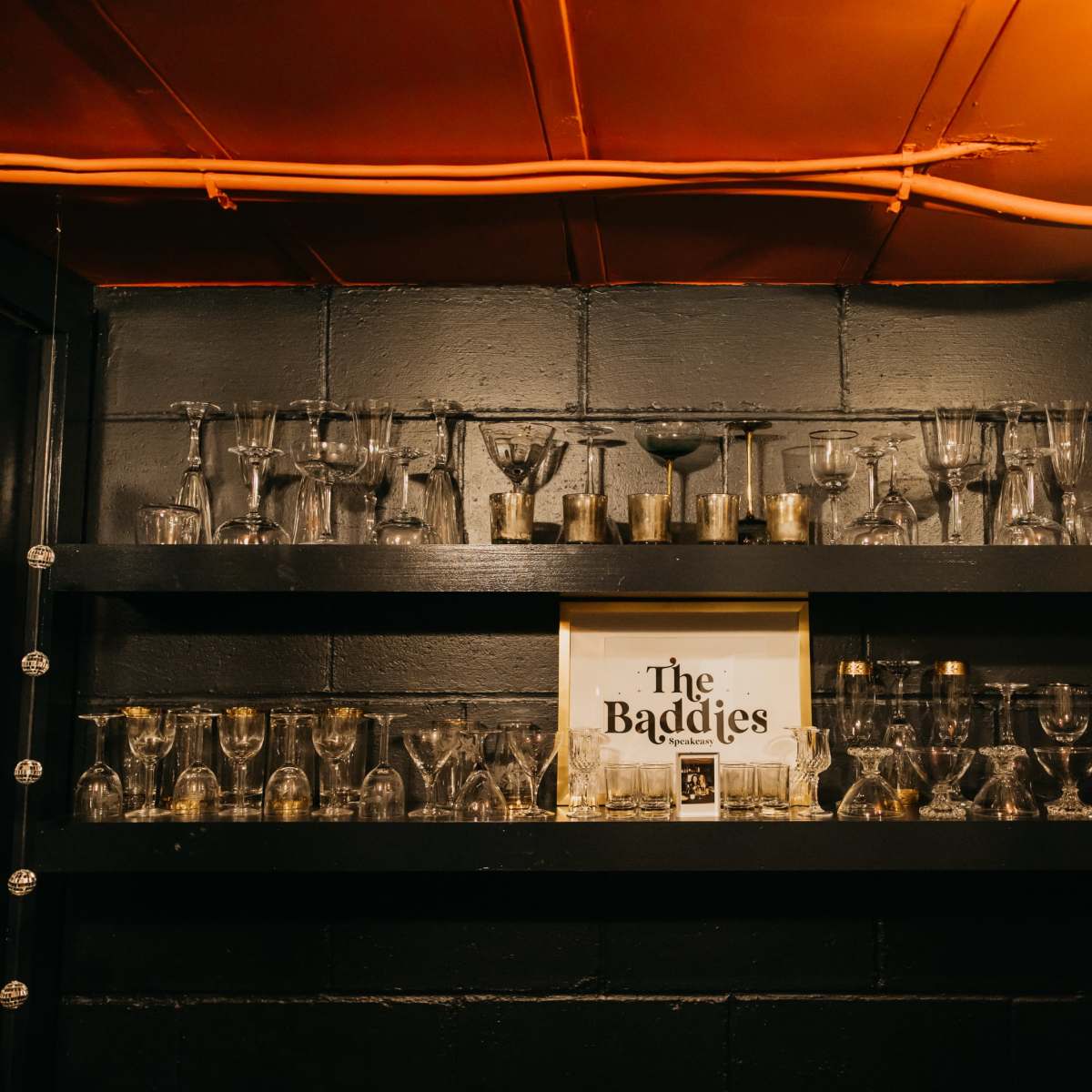 A black wall shelf displays various glassware and a framed sign that reads "The Baddies."