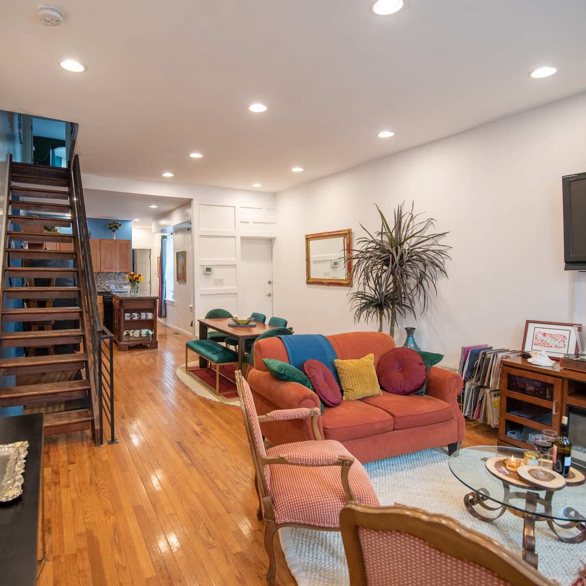 A bright living room with a staircase leading to the upper level, featuring orange and green furnishings and wooden flooring.
