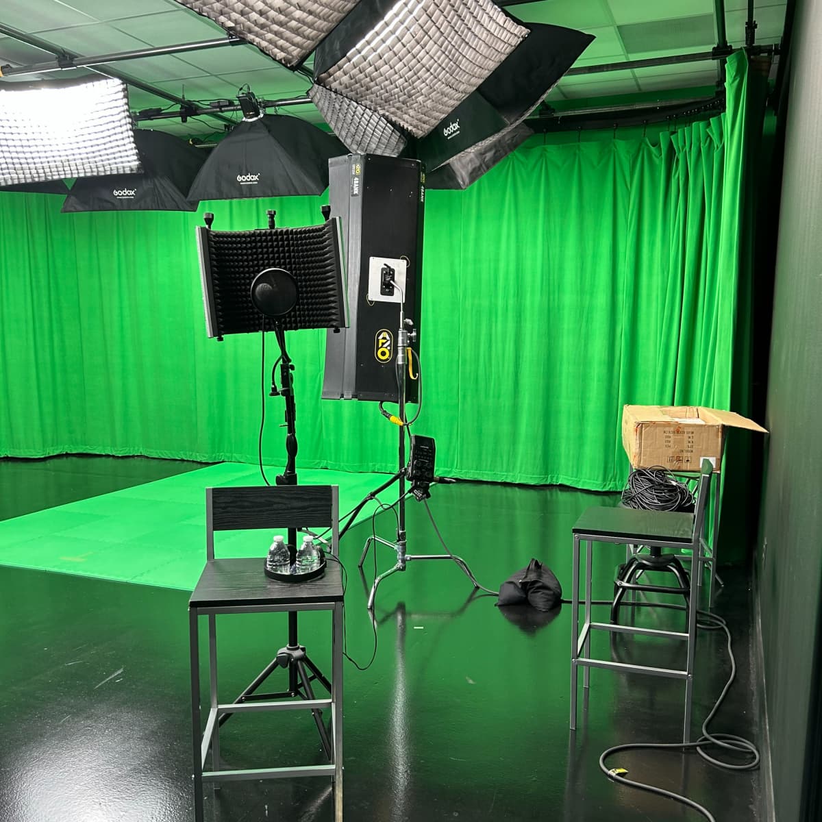 A green screen studio setup with lighting equipment, two empty chairs, and cables on a glossy black floor.