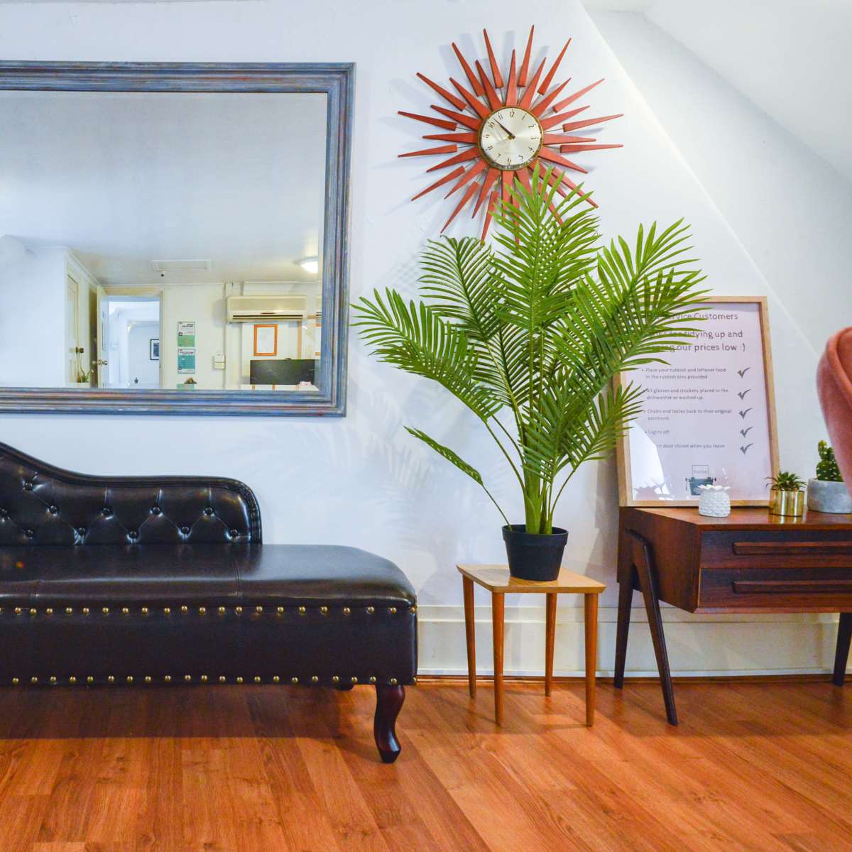 A black tufted chaise lounge positioned near a large mirror, a potted plant, and a wooden side table in a well-lit interior space.