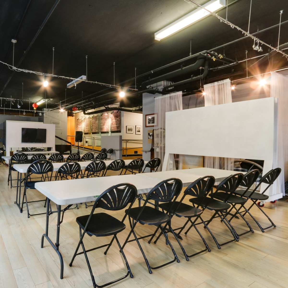 A spacious room set up with several rows of folding chairs facing a series of tables, with a projector screen and artworks displayed on the walls.