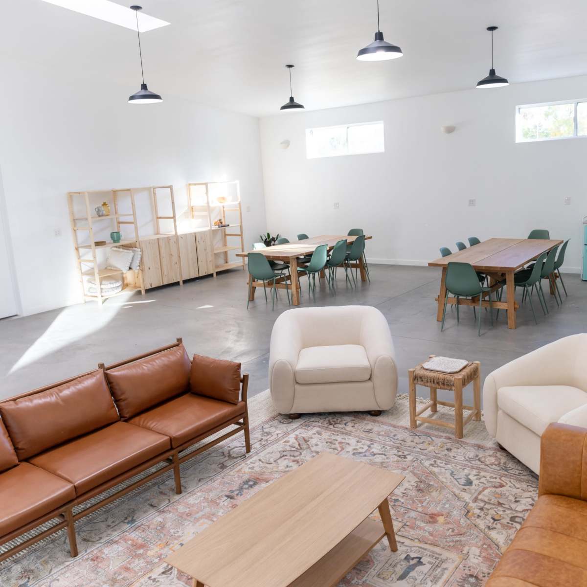 A spacious, modern room with a seating area featuring brown and cream sofas, a wooden coffee table, and a dining area with green chairs and a wooden table under pendant lighting.