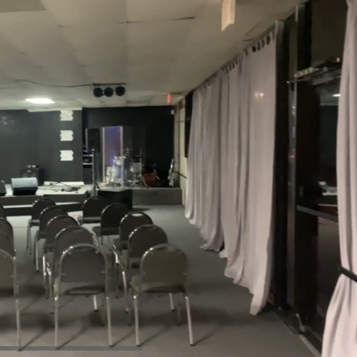 A row of empty chairs faces a stage in a dimly lit, spacious room with musical instruments in the background.