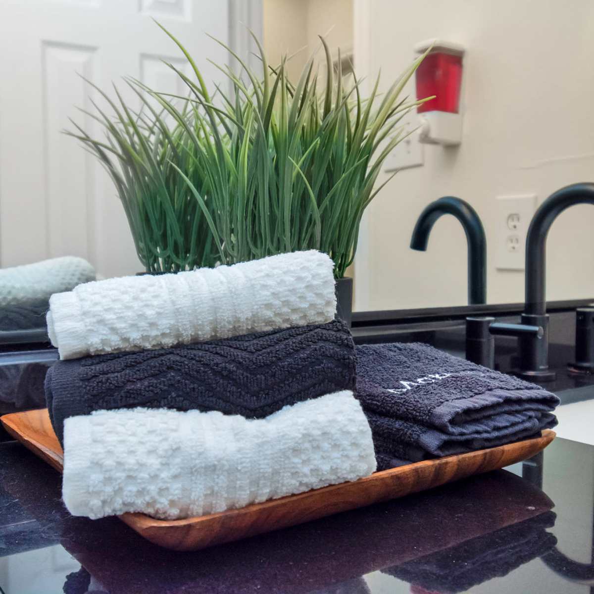 A wooden tray holds neatly stacked black and white towels next to a small potted plant in a bathroom setting with a sink and faucet visible in the background.