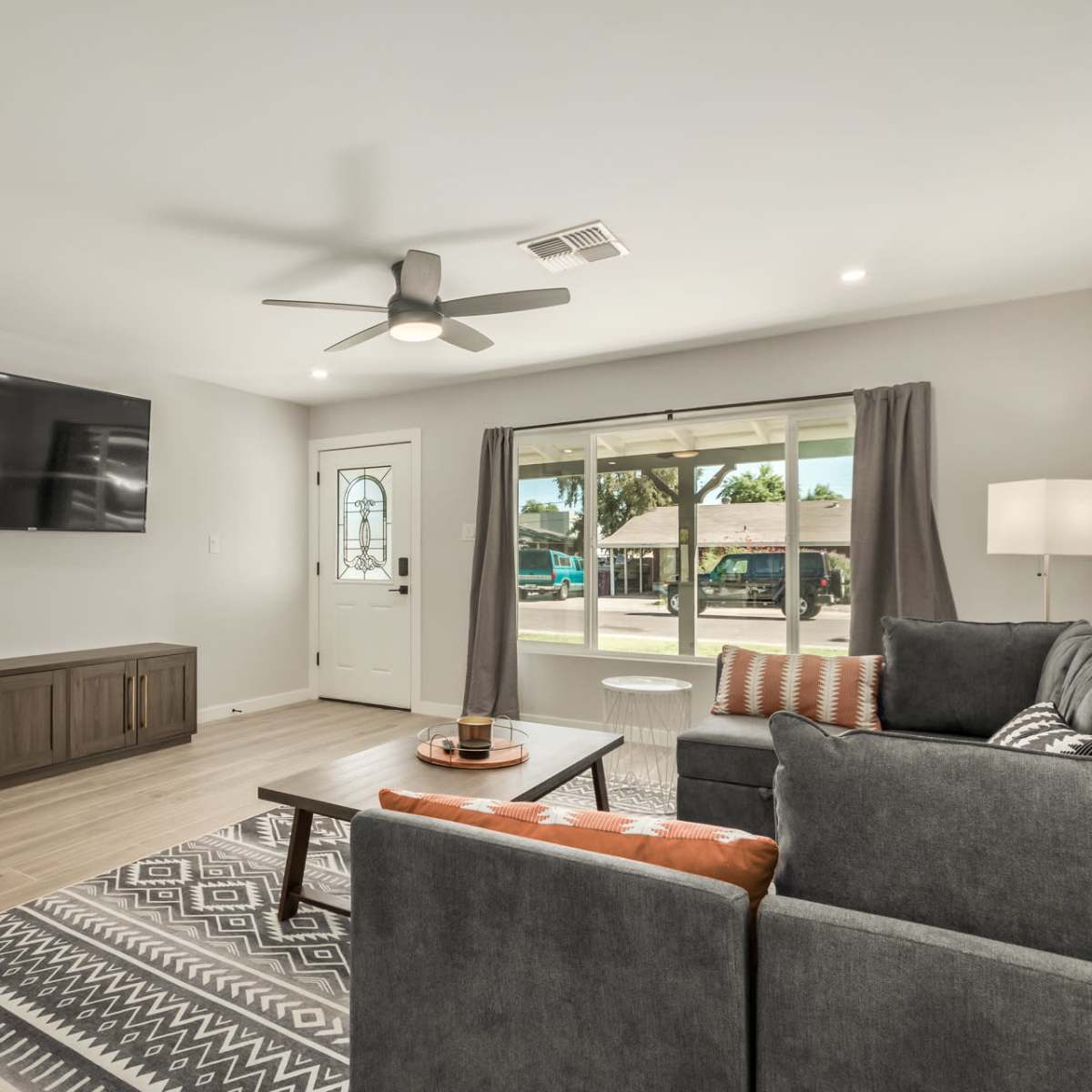 A modern living room features a gray sectional sofa, a coffee table, a wall-mounted television, and a large window with natural light pouring in.