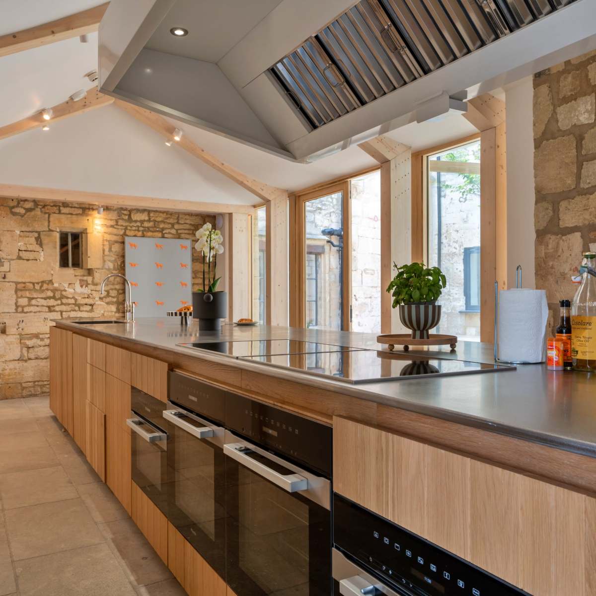 The kitchen features a central island with built-in appliances surrounded by stone walls and large windows that provide natural light.