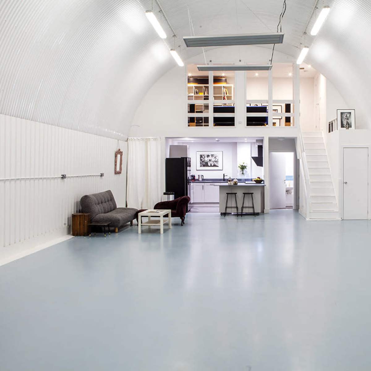 A spacious, modern interior with high arching white walls, a light blue floor, and a loft area featuring a kitchen and dining space.