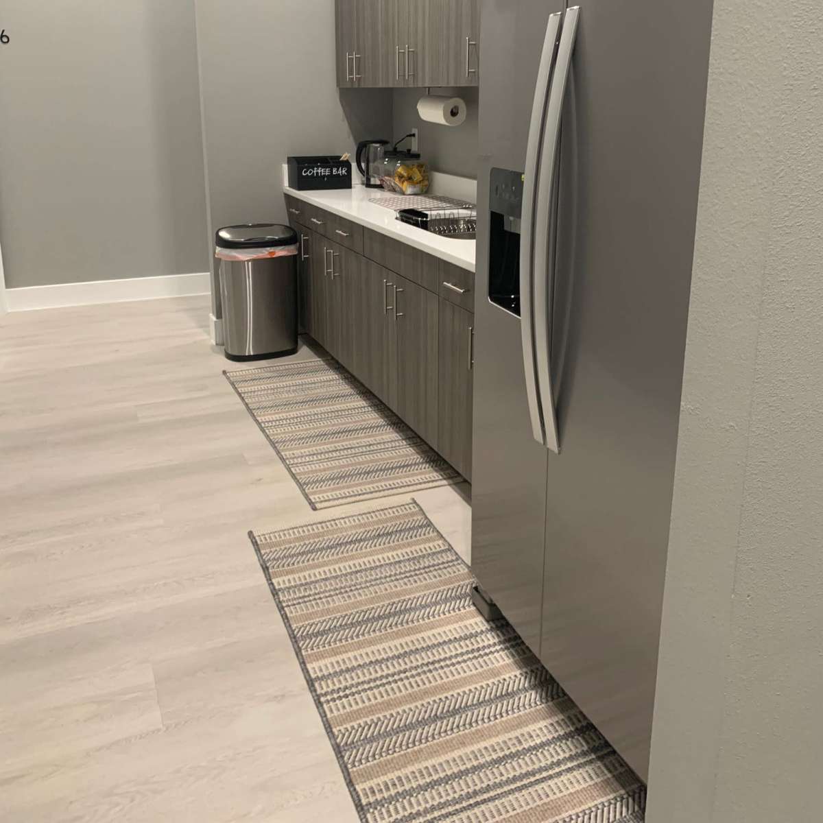 A modern kitchen featuring a stainless steel refrigerator, wooden cabinets, a countertop with kitchen appliances, and a trash can.