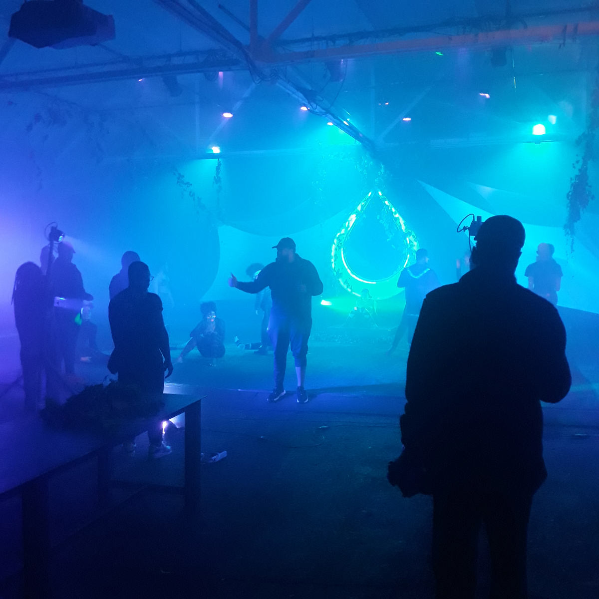 A group of people gathers in a dimly lit space with blue and green lighting, surrounded by fog and decorative elements.