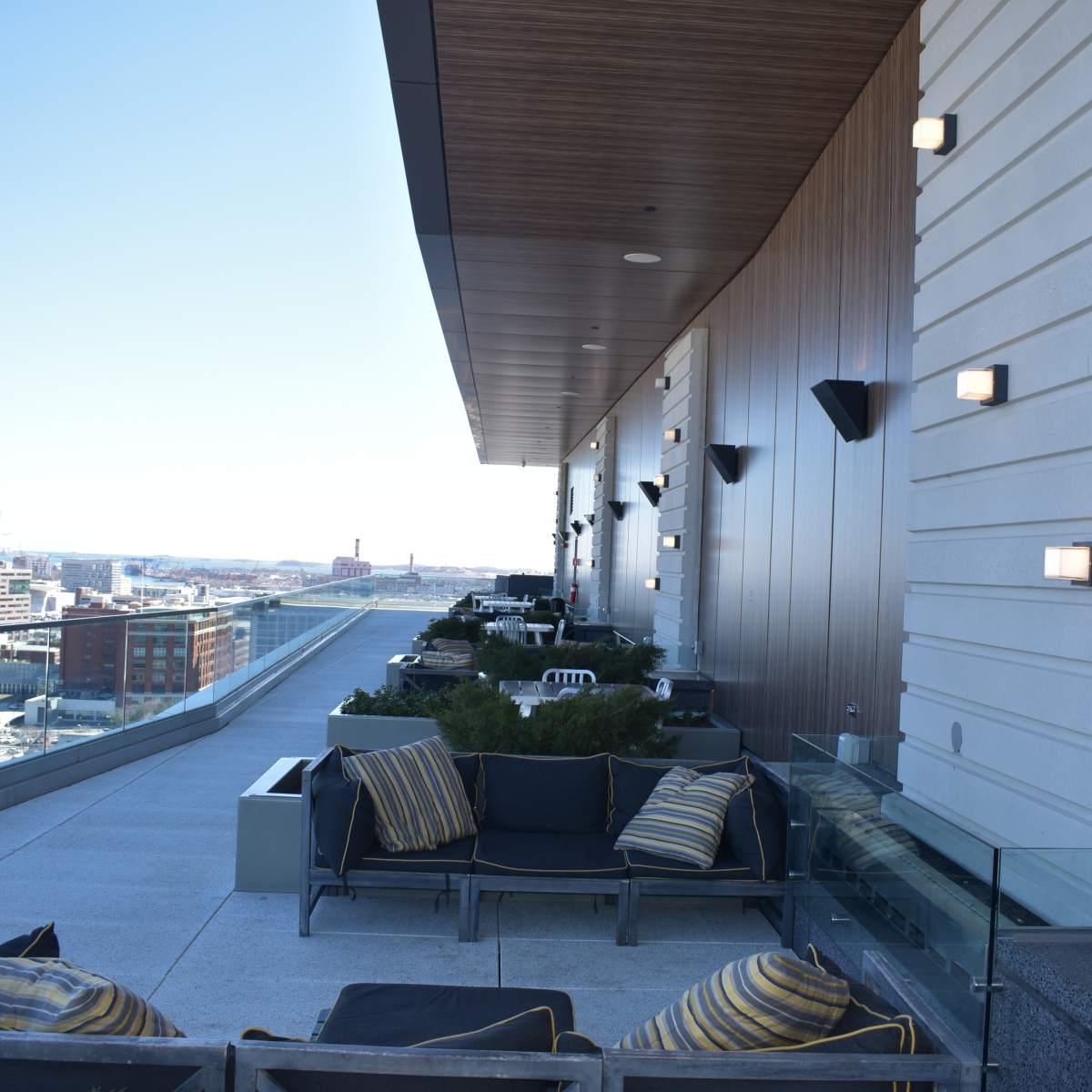A modern rooftop terrace with seating areas, planters, and wall-mounted lights, overlooking a city skyline.