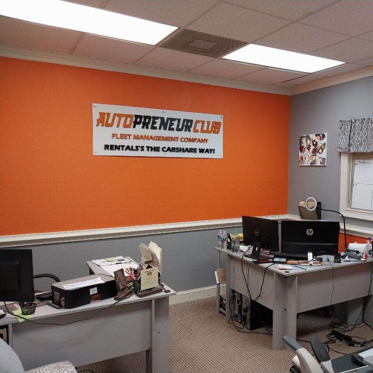 An office space with orange and gray walls, featuring a desk with a computer and office supplies, along with a large banner for "AutoPreneur Club" on the wall.