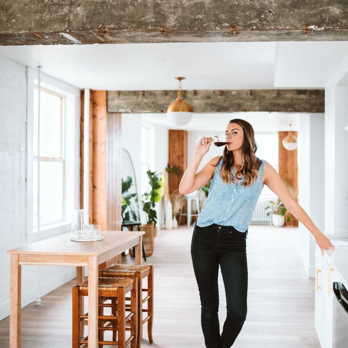 A woman stands in a modern kitchen, sipping from a wine glass while leaning against a white countertop.