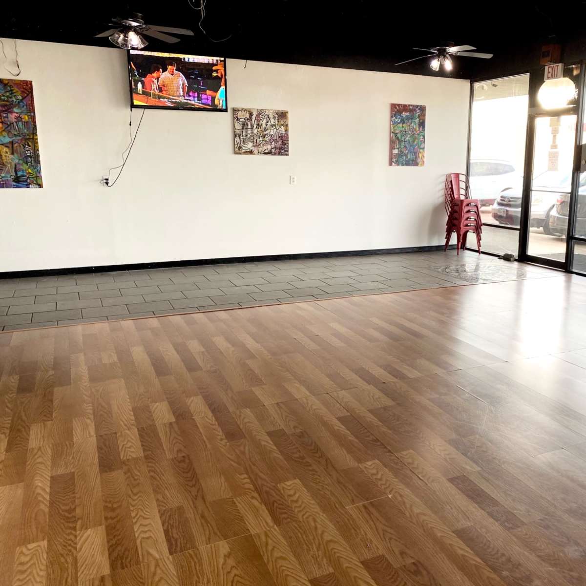 A spacious room with wood and tile flooring, a television mounted on the wall, and large windows displaying parked cars outside.