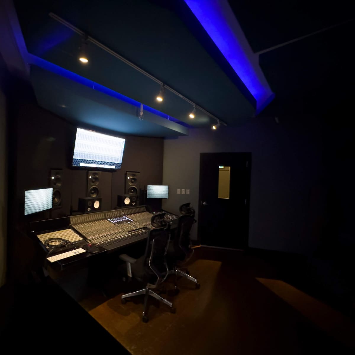 A recording studio with a control desk, speakers, and two office chairs in a dimly lit space.
