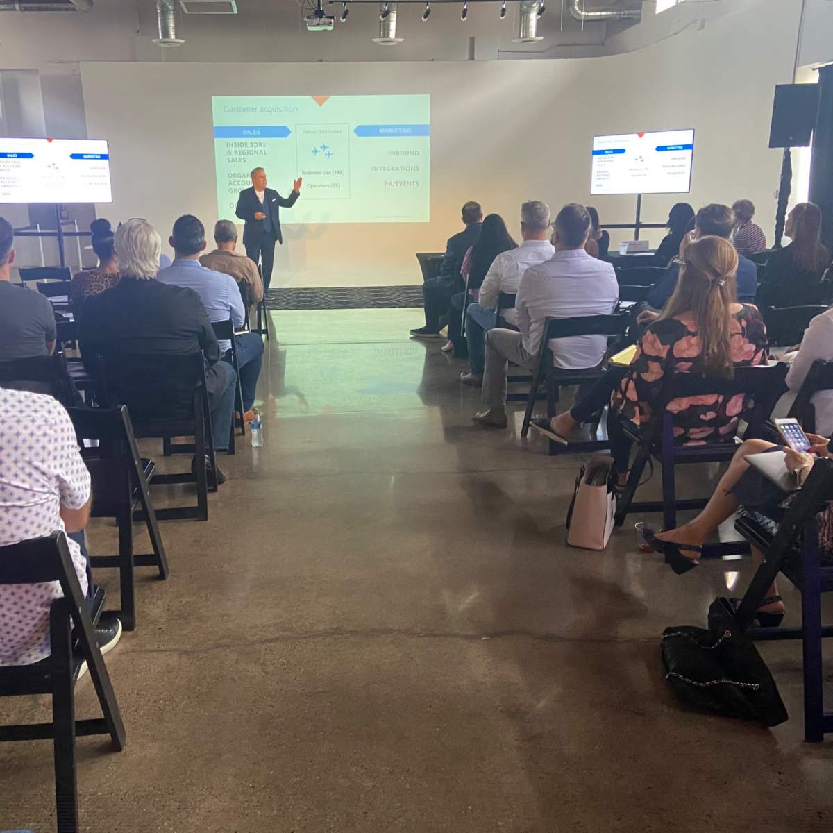 A speaker presents to an audience seated in a modern conference space with a screen displaying information.