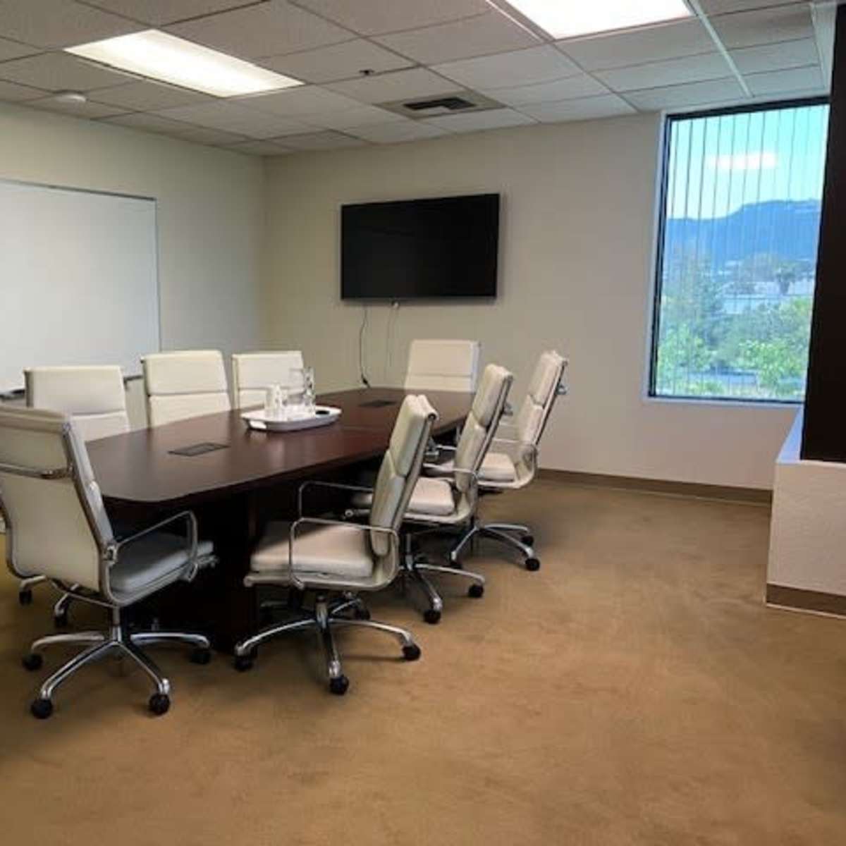 A conference room features a large wooden table surrounded by eight white leather chairs, with a wall-mounted television and a whiteboard visible in the background.