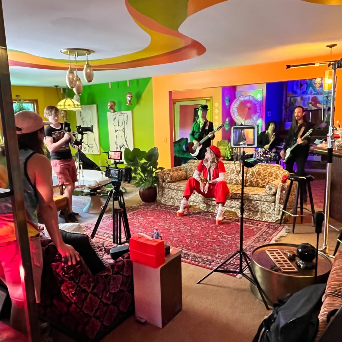 A group of people is filming a music video in a brightly colored room with a vintage sofa and various sets of lights and cameras.