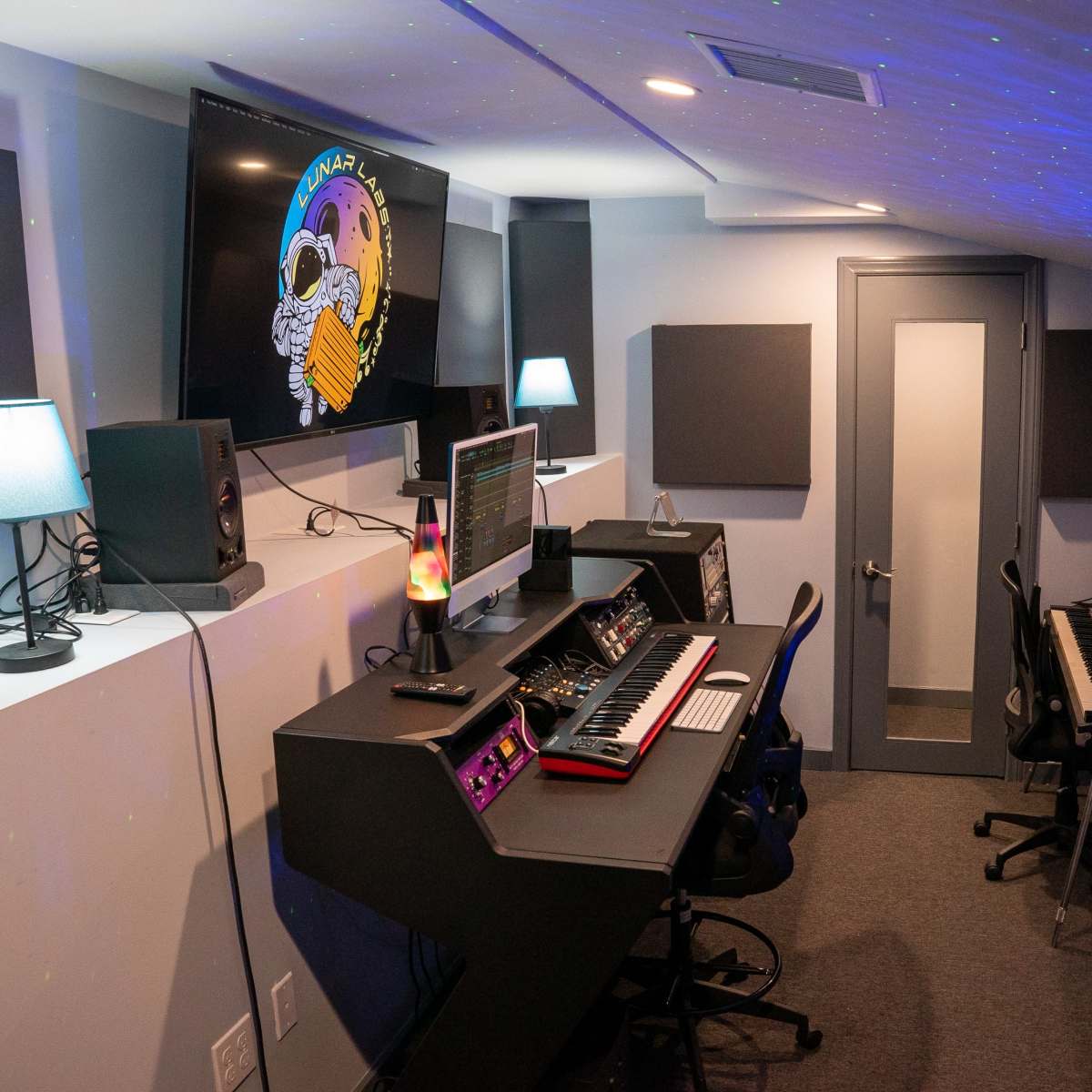 A music studio equipped with a computer, synthesizers, speakers, and a keyboard, featuring a glowing lamp and soundproof panels on the walls.