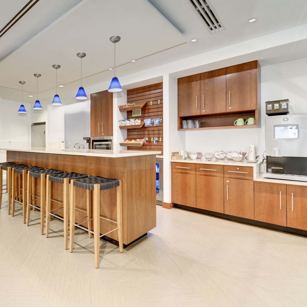 A modern kitchen area features a long island with bar stools, overhead lights, and a coffee station against a wall of cabinets.