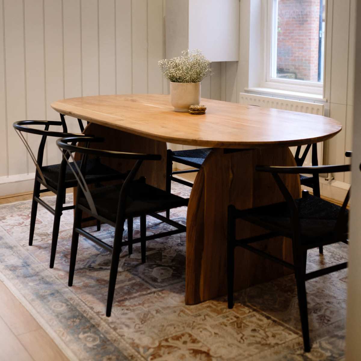 A wooden oval dining table surrounded by black chairs sits under three pendant lights in a well-lit room with white paneling.