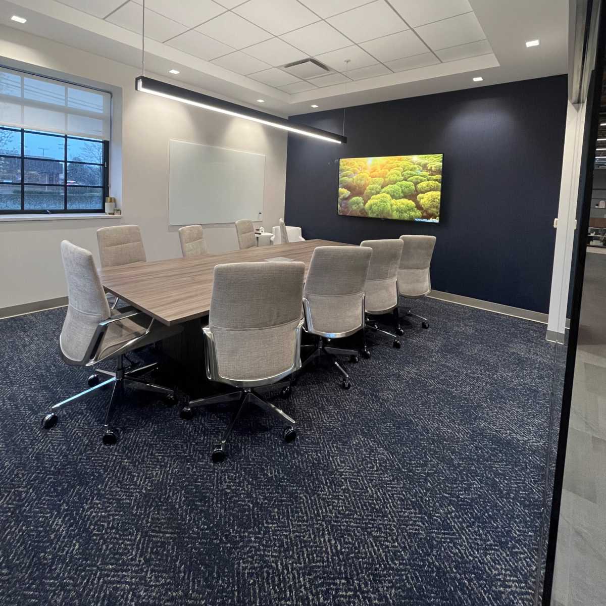 A modern conference room features a large table surrounded by ergonomic chairs, a whiteboard, and a wall-mounted screen displaying a colorful image.