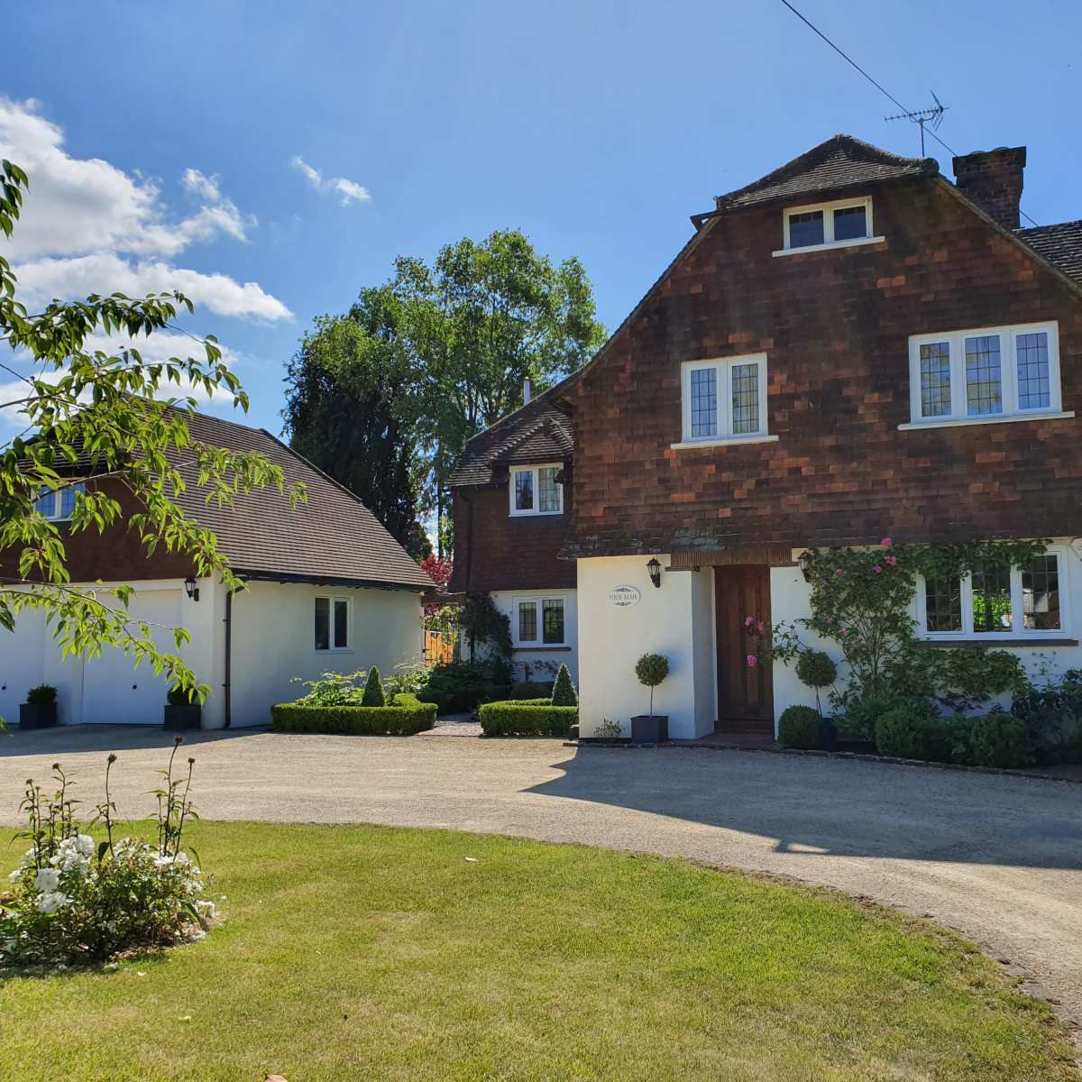 A two-story house with a distinctive wooden shingle facade, surrounded by green lawns and trees, along with a gravel driveway leading up to it.
