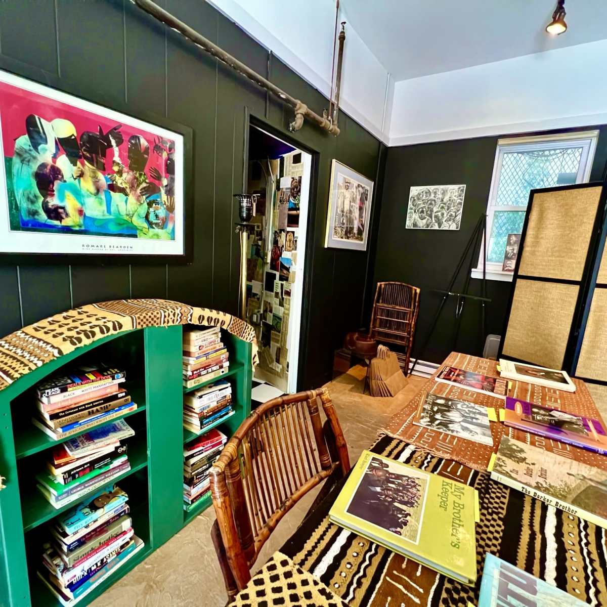 A cozy room with green bookshelves filled with books, a wooden table displaying various publications, and artwork adorning the dark walls.