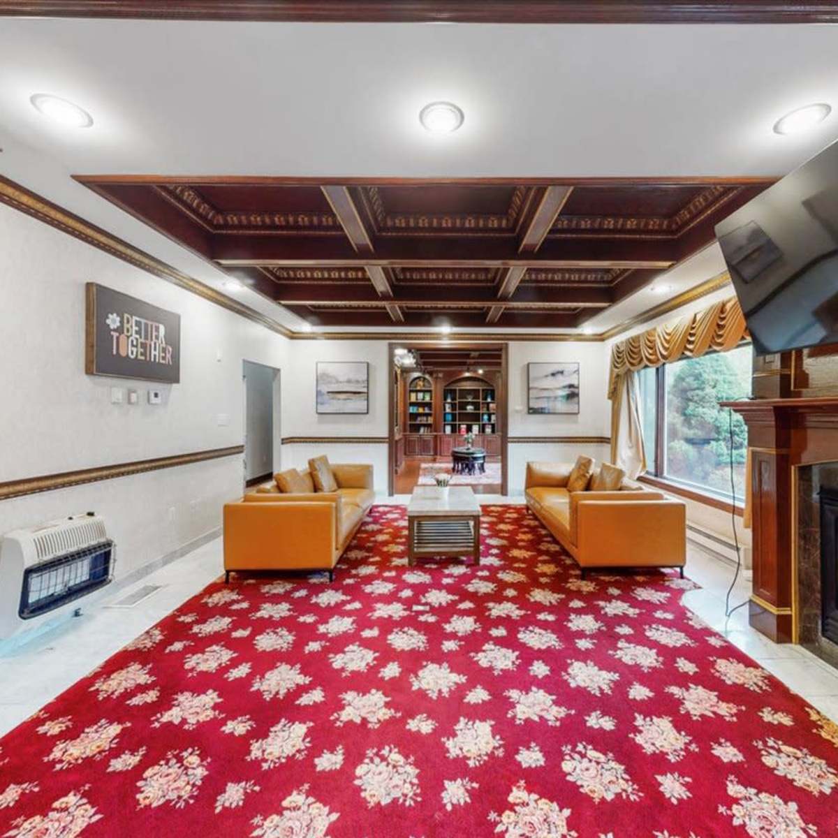 A cozy living room with orange sofas arranged around a coffee table, a large TV mounted on the wall, and a decorative red carpet.