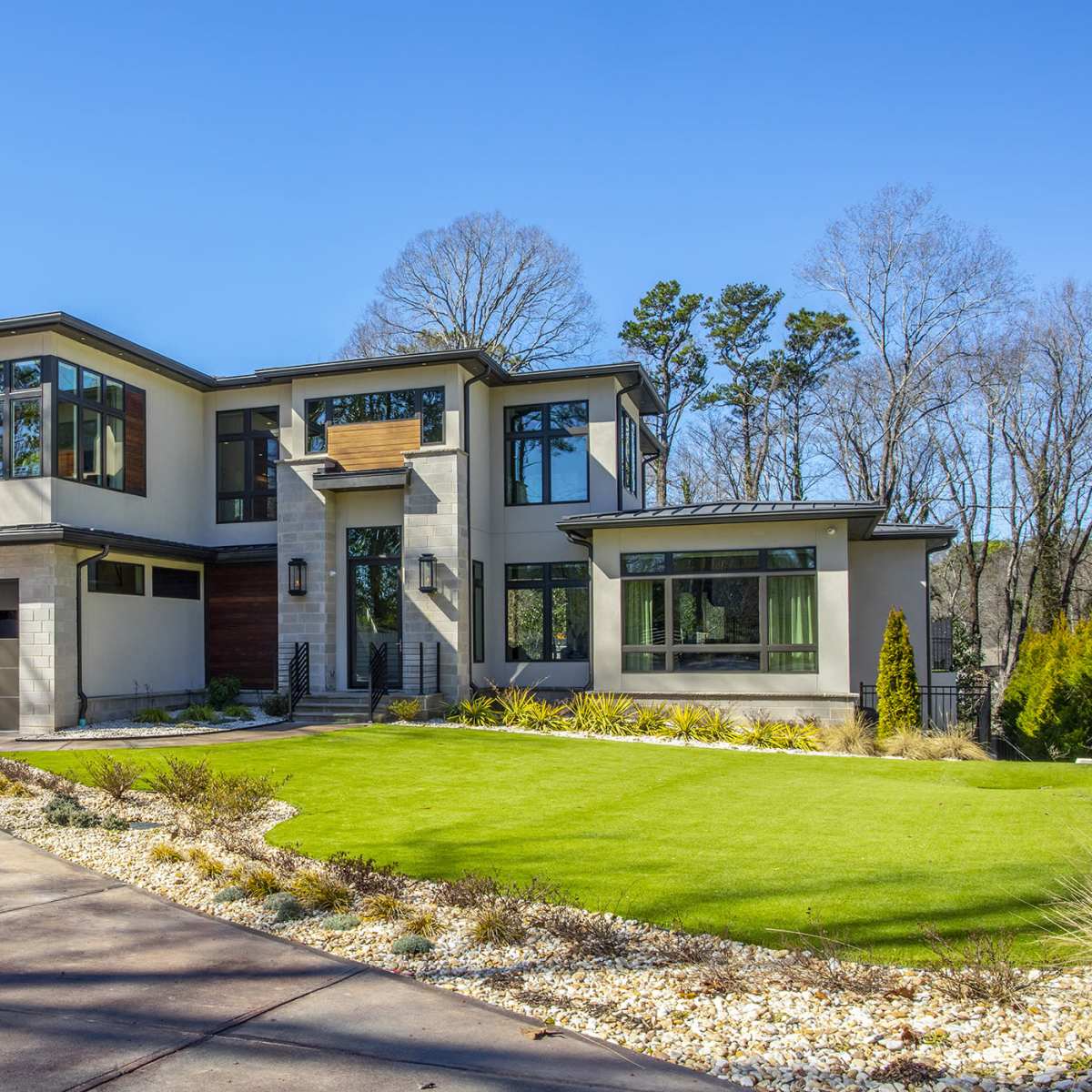 A modern two-story house features large windows, a stone and wood facade, and a well-manicured lawn with landscaped gardens.