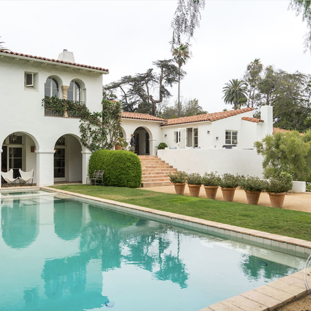 A two-story white stucco house with arched windows and a pool in the foreground, surrounded by manicured greenery and potted plants.