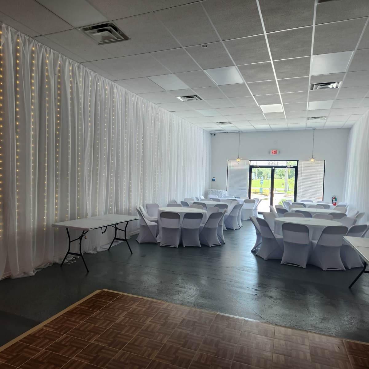 A decorated event space with white draped curtains, string lights, and tables arranged with white covers.
