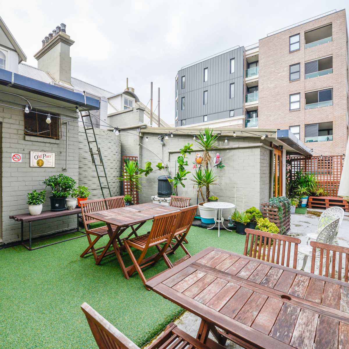 A patio area with wooden tables and chairs, surrounded by potted plants, against a backdrop of both a traditional and modern building.