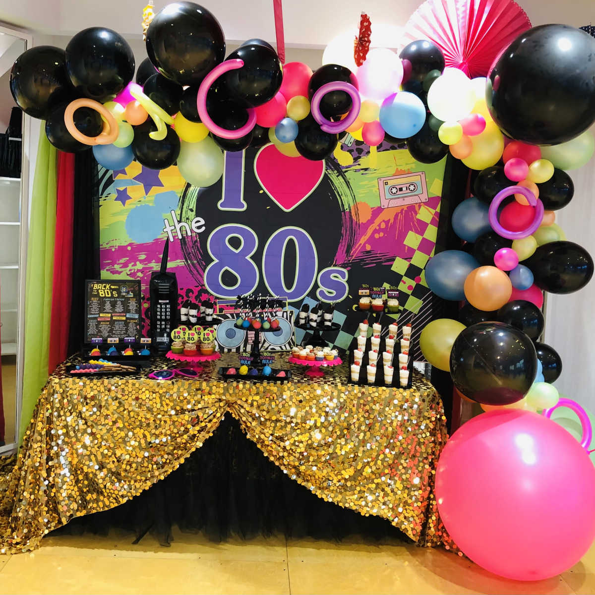 A decorated table for an 80s-themed party with a colorful backdrop, black and pink balloons, and a variety of treats arranged on a shiny gold tablecloth.