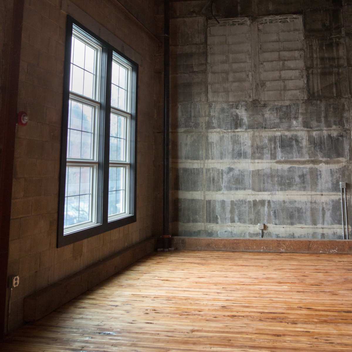 An interior space with exposed wooden floors and a large window on a painted wall, adjacent to an unfinished concrete wall.
