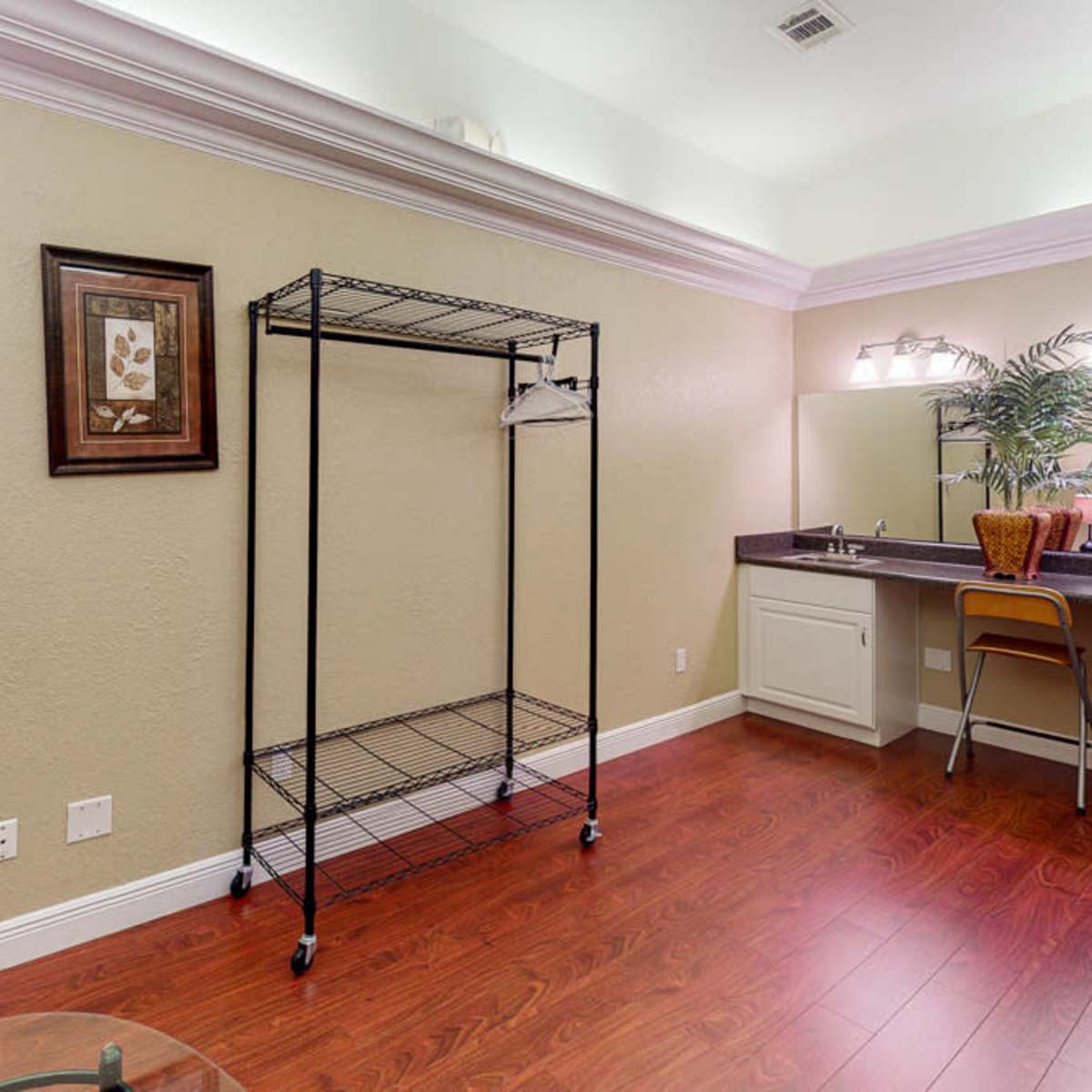 An interior room with a wire clothing rack, a desk with a chair, and a mirror, all set against a light-colored wall and hardwood floor.