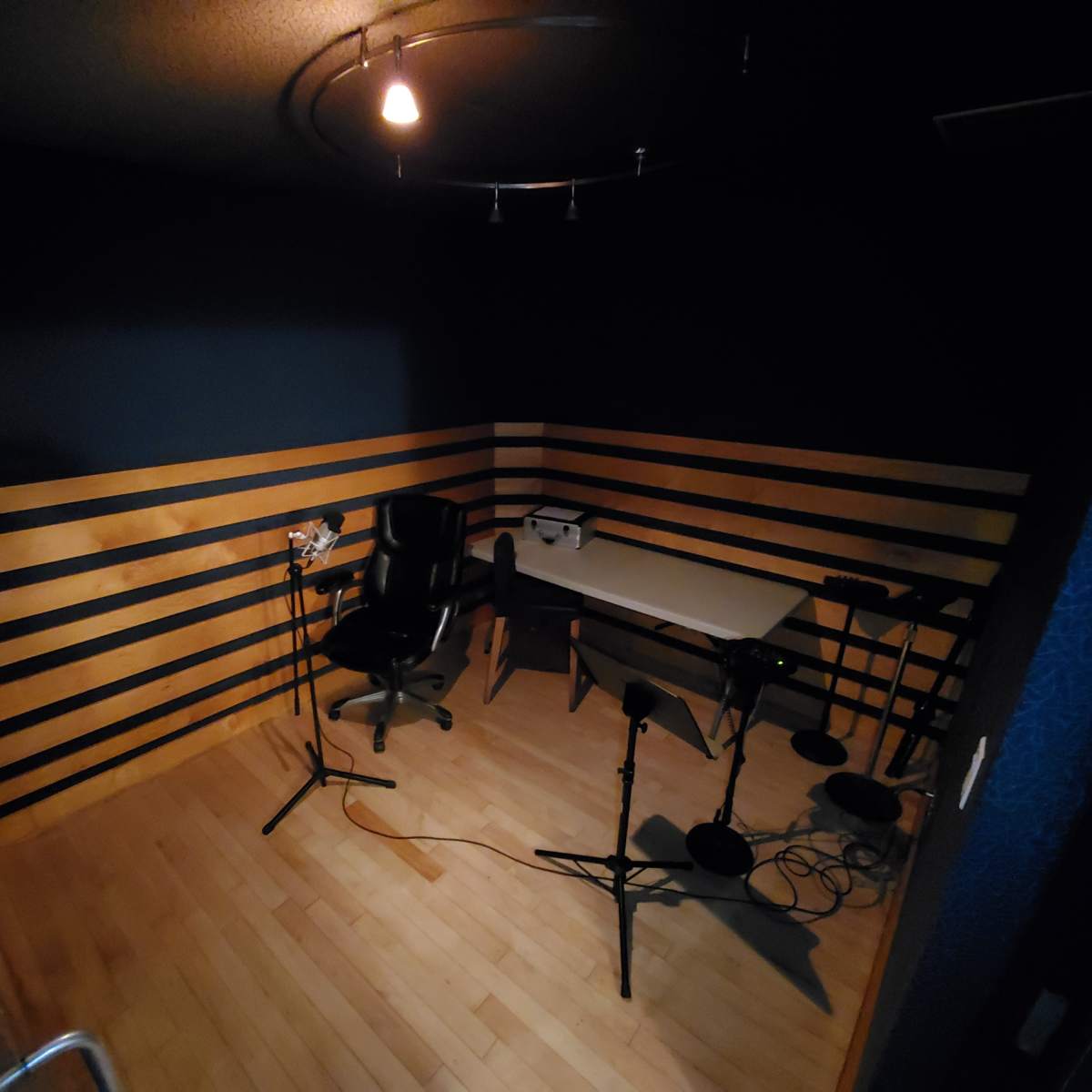 A small recording studio featuring wooden striped walls, a black office chair, a table, and microphones on stands.