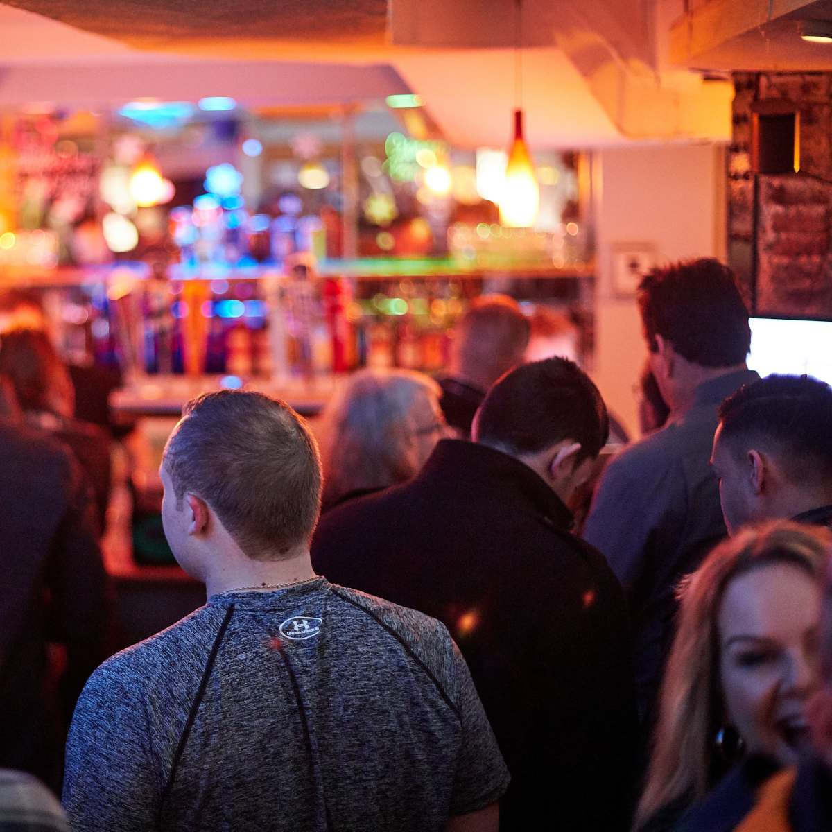 A group of people stands with their backs to the camera in a dimly lit bar, with a brightly lit bar counter visible in the background.