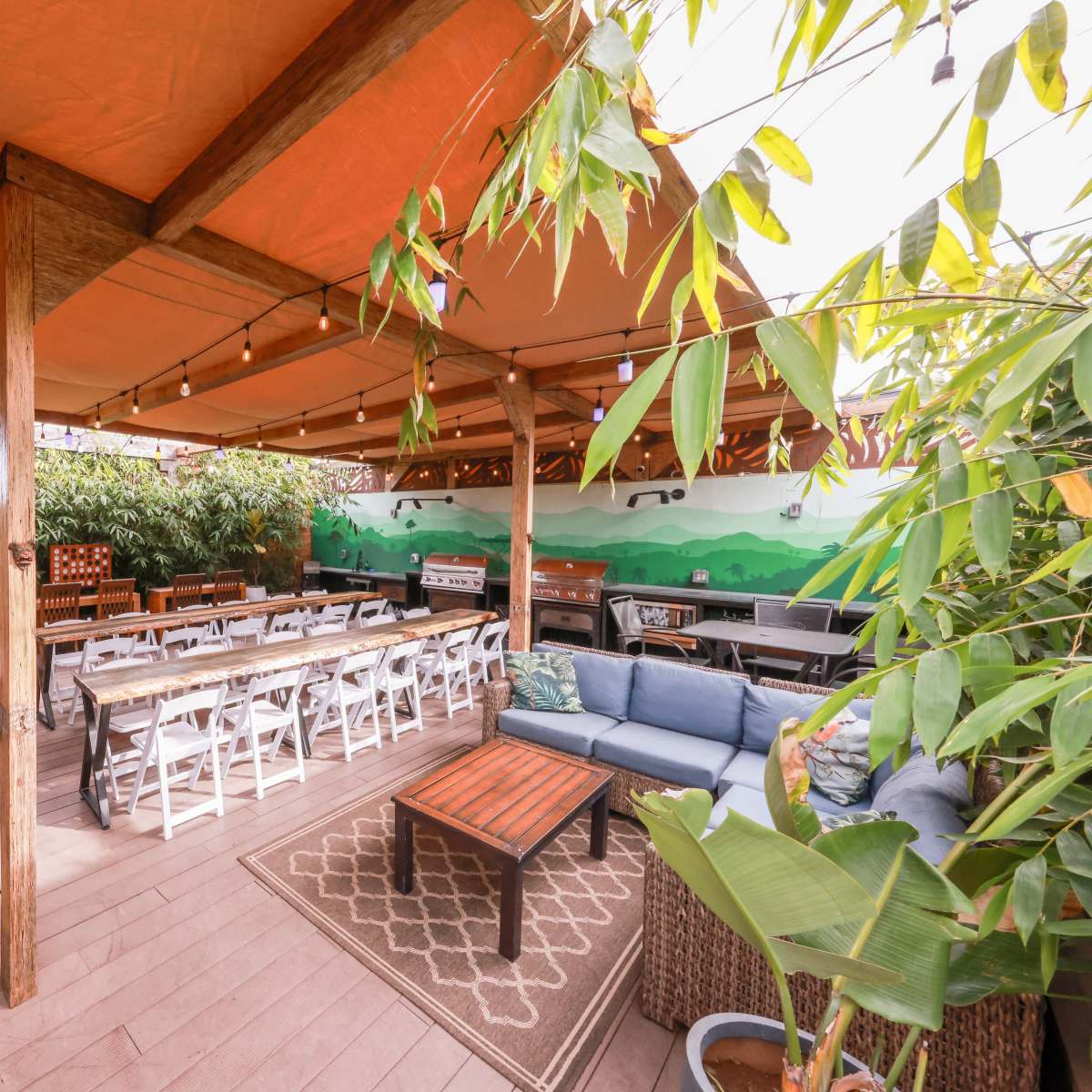 A spacious outdoor seating area with wooden tables, white chairs, and a large sofa, all under a canopy of orange fabric and surrounded by greenery.