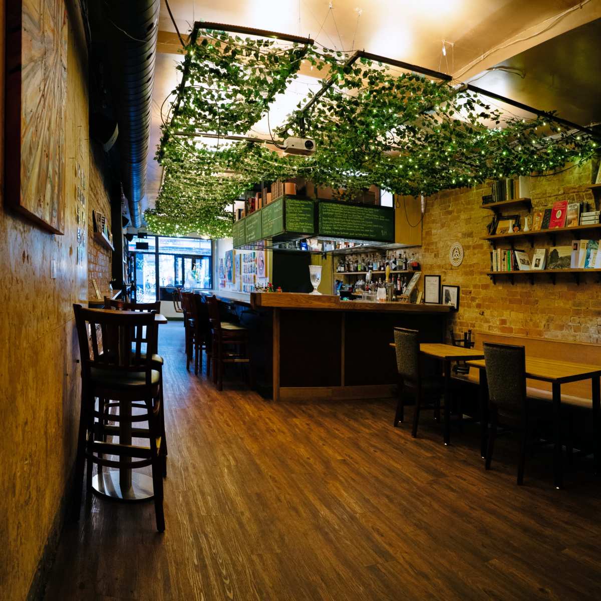 A cozy café interior with wooden flooring, a bar area, and shelves filled with books, complemented by greenery hanging from the ceiling.