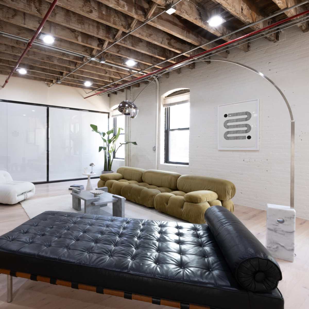 A spacious, modern living room with exposed wooden beams, a green sofa, a black chaise lounge, and large windows allowing natural light.