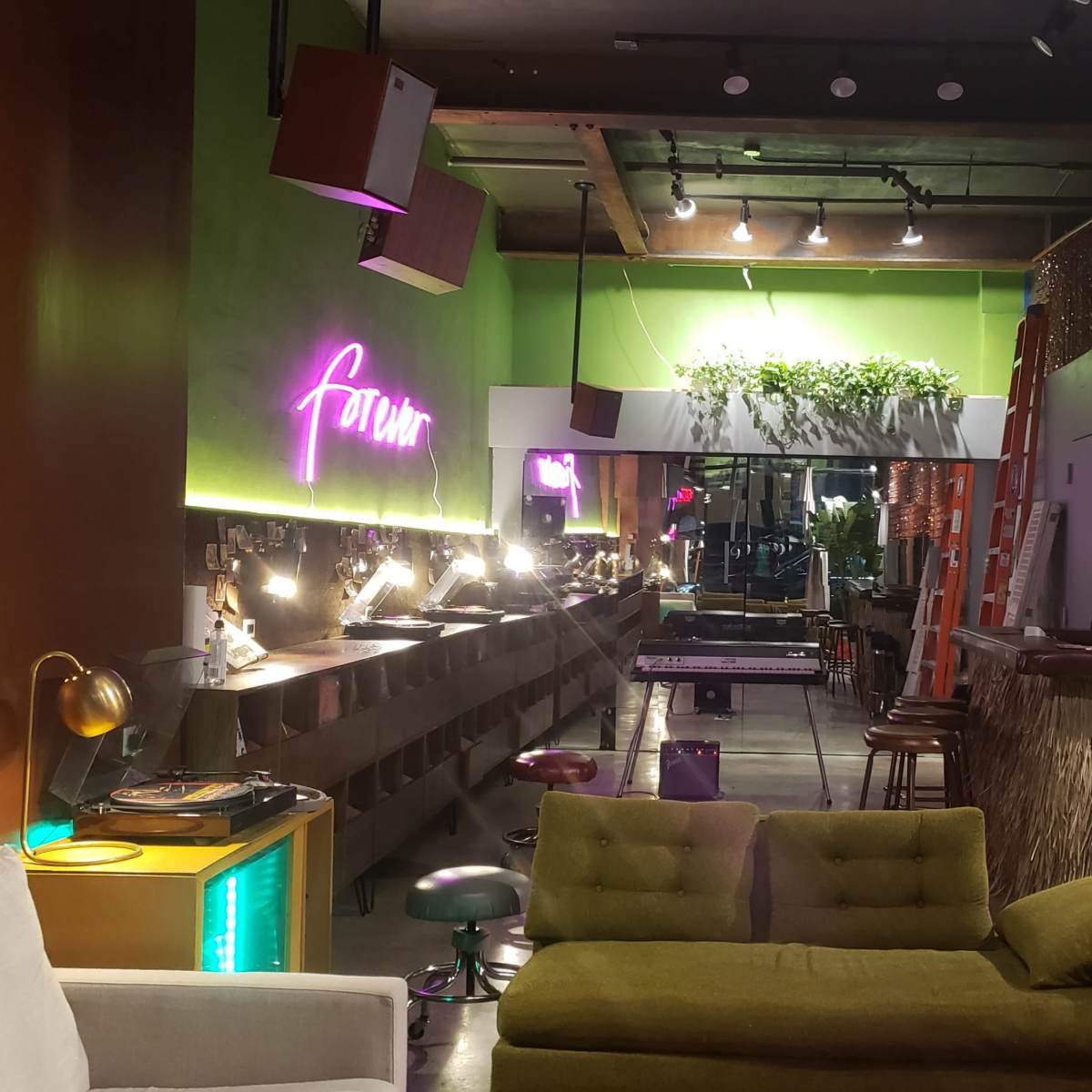 A brightly lit bar interior features vibrant green walls, a neon "forum" sign, and various seating arrangements alongside a long counter with bar tools and decor.