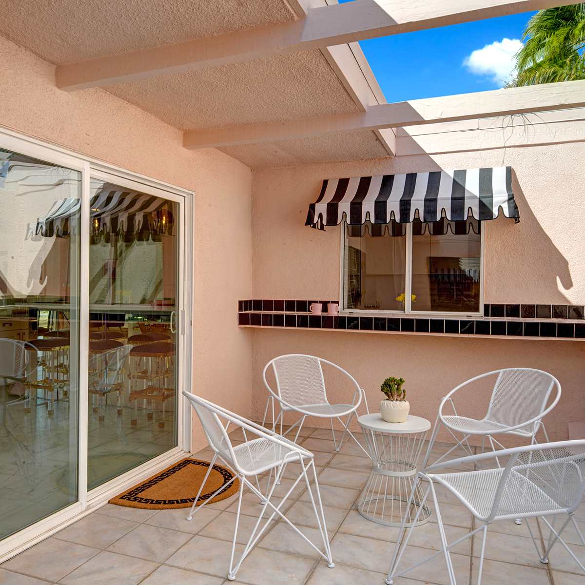 A patio with white metal chairs and a round table, featuring large glass doors and a window with a striped awning.